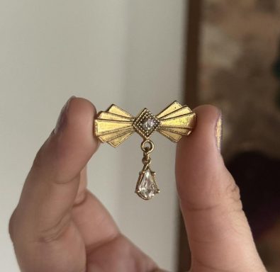 Gold toned geometric bow brooch with a rhinestone in the center and a hanging teardrop crystal.