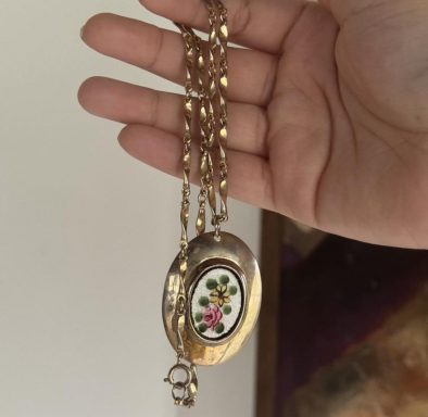 Gold toned oval pendant with a pearly painting in the center featuring a yellow and pink flower. Pendant hangs on a gold twisted chain.